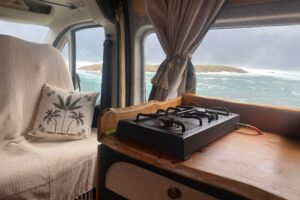 Blick auf einen Gaskocher im Campervan, links ein Kissen mit einer Palme, draußen das tobende Meer