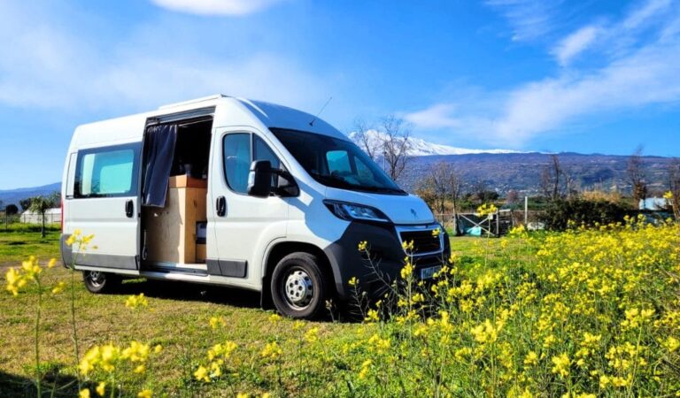 Ein weißer Campervan steht in einer gelben Blumenwiese, im Hintergrund der Ätna auf Sizilien