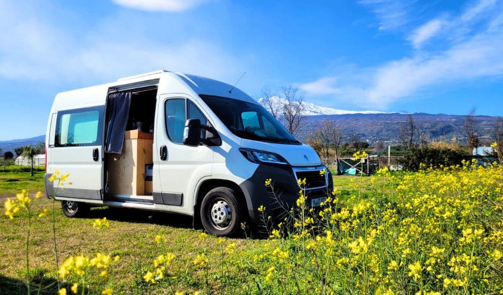 Ein weißer Campervan steht in einer gelben Blumenwiese, im Hintergrund der Ätna auf Sizilien
