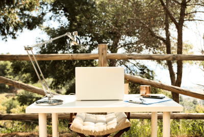 Ein Outdoor-Arbeitsplatz bestehend aus einem hellen Tisch mit weißem Laptop, einer Tasse und einer Lampe darauf, steht draußen im Grünen